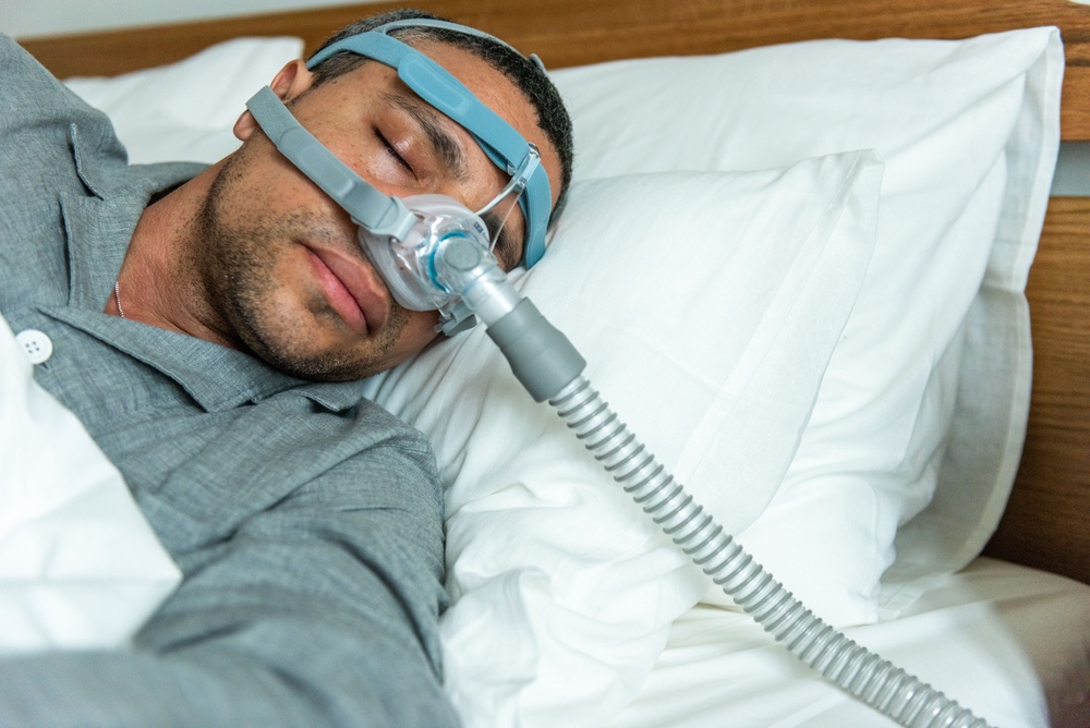 A person sleeps in bed wearing a CPAP mask for sleep apnea therapy. The setting is a cozy bedroom with white pillows and sheets, creating a restful atmosphere.