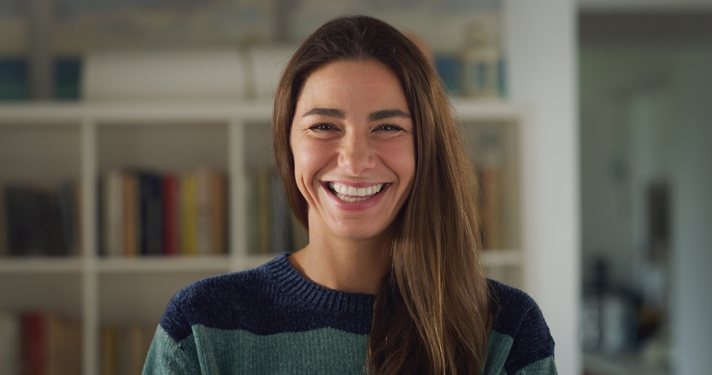 Cinematic shot of attractive happy young woman with colorful sweater is smiling in camera in living room at home. Concept of life, family, health, happiness, femininity, women's day.