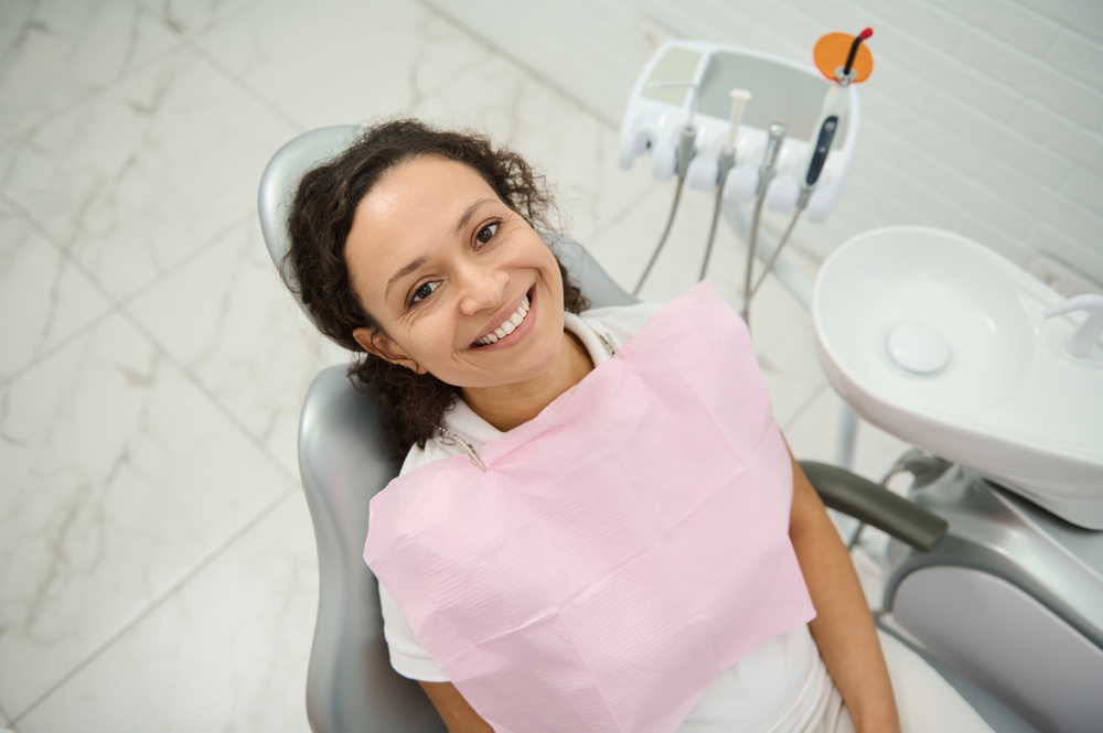 High angle view charming dark-haired woman smiles with beautiful healthy toothy smile looking at camera while sitting in dentists chair during regular dental check-up at dentist hygienist appointment