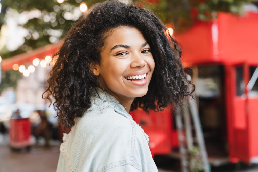Cosmetic dentistry smile confidence in the Williamsburg community Young woman with curly hair smiling over her shoulder outdoors, confident smile from cosmetic dentistry in the Williamsburg community