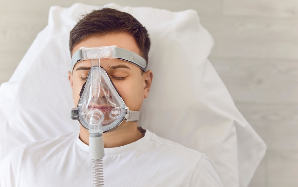 Young adult man lying on a hospital couch, sleeping and breathing through an oxygen mask. Medical treatment supports healthcare needs, addressing asthma or lung therapy requirements.