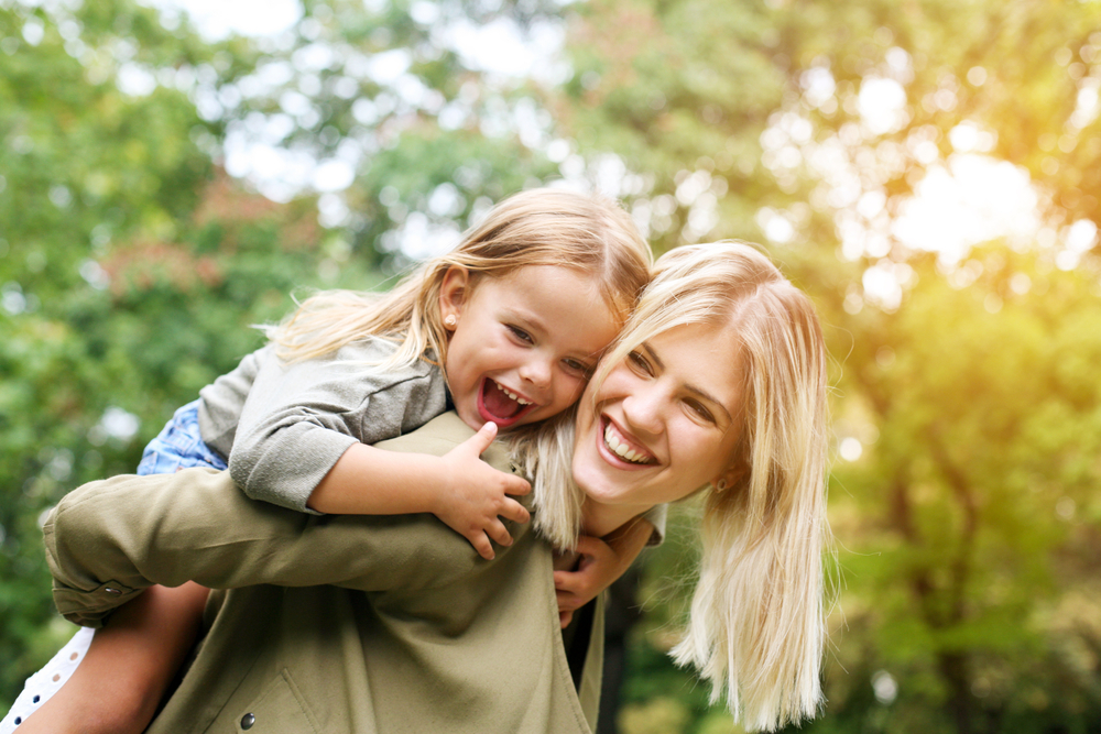Mother and Daughter Smiling: Virginia Beach Community Whitening Patient Mother giving her daughter a piggyback ride outdoors after a teeth whitening visit in the Virginia Beach community