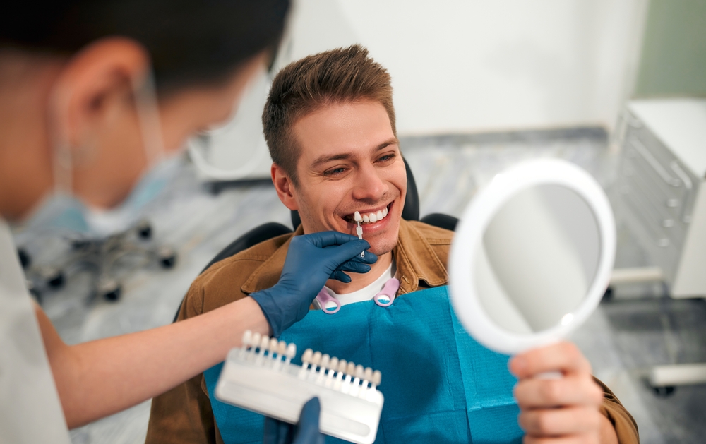 Male patient reviewing his smile in a mirror while a dentist matches veneer shading at The Foleck Center, Virginia Beach, VA 