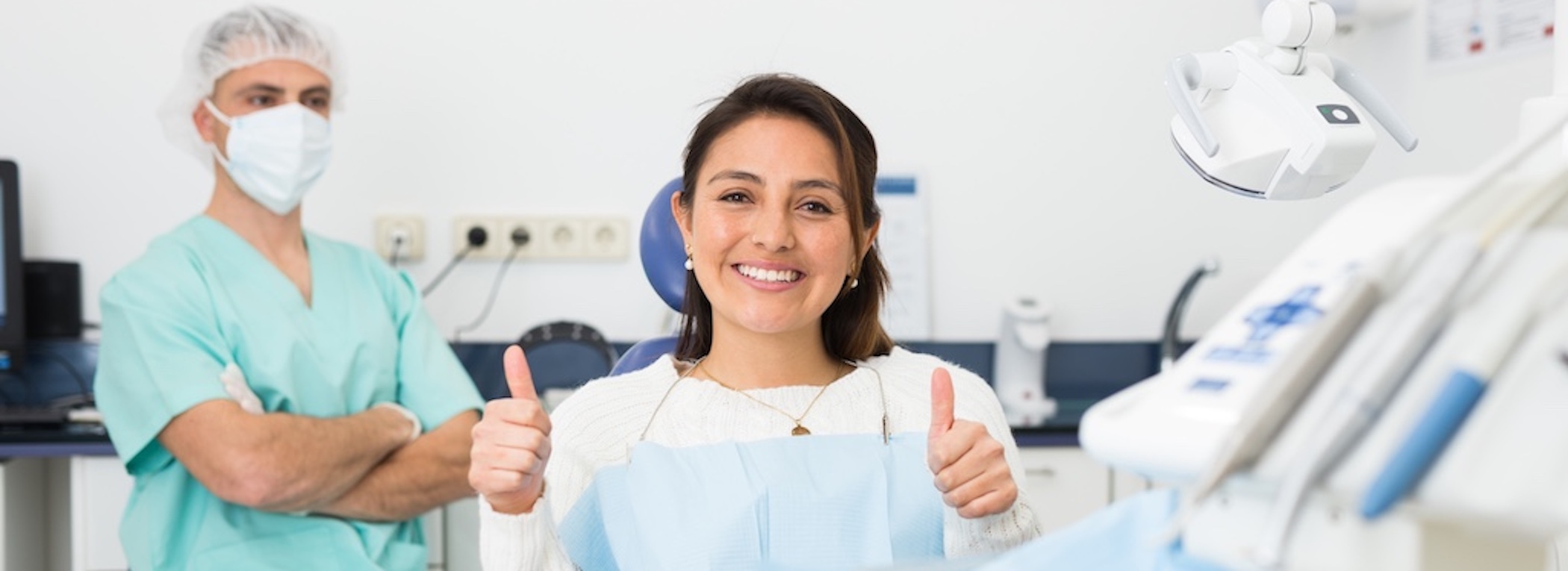 Happy Latina woman sitting in a dental chair after a full-mouth dental implants procedure, giving a thumbs up.