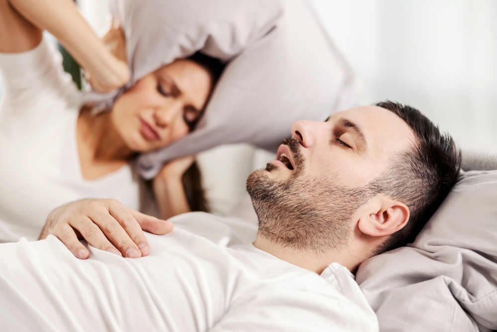 Woman covering her ears with a pillow while her partner snores, illustrating a common sign of sleep apnea in Hampton 