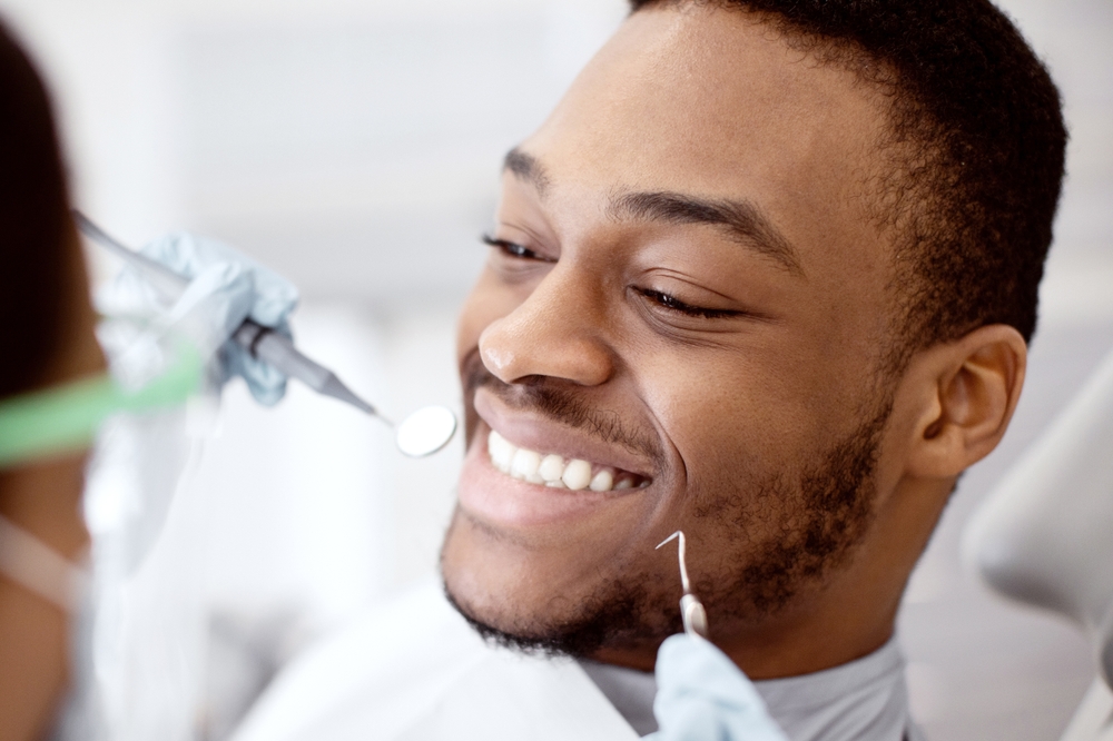 Comprehensive dental checkups in Hampton Roads Young man smiling during a dental exam as a dentist uses a mirror and explorer tool
