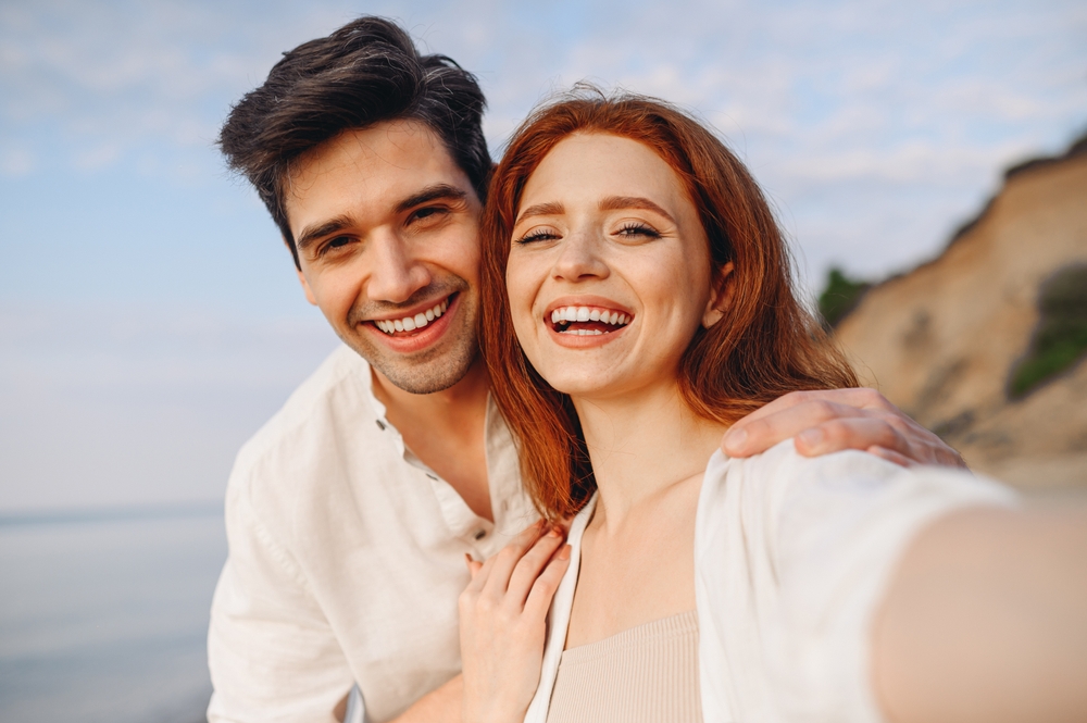 Custom dental restorations in Virginia Beach, VA, The Foleck Center Smiling couple taking a selfie at the beach with bright, healthy teeth, reflecting confidence after dental lab restorations at The Foleck Center, Virginia Beach, VA