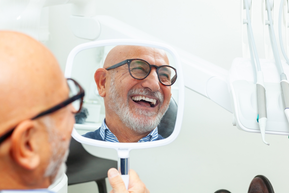 Dentures and partials results in Virginia Beach, VA, The Foleck Center Older man smiling confidently at his new smile in a dental hand mirror after denture treatment at The Foleck Center, Virginia Beach, VA
